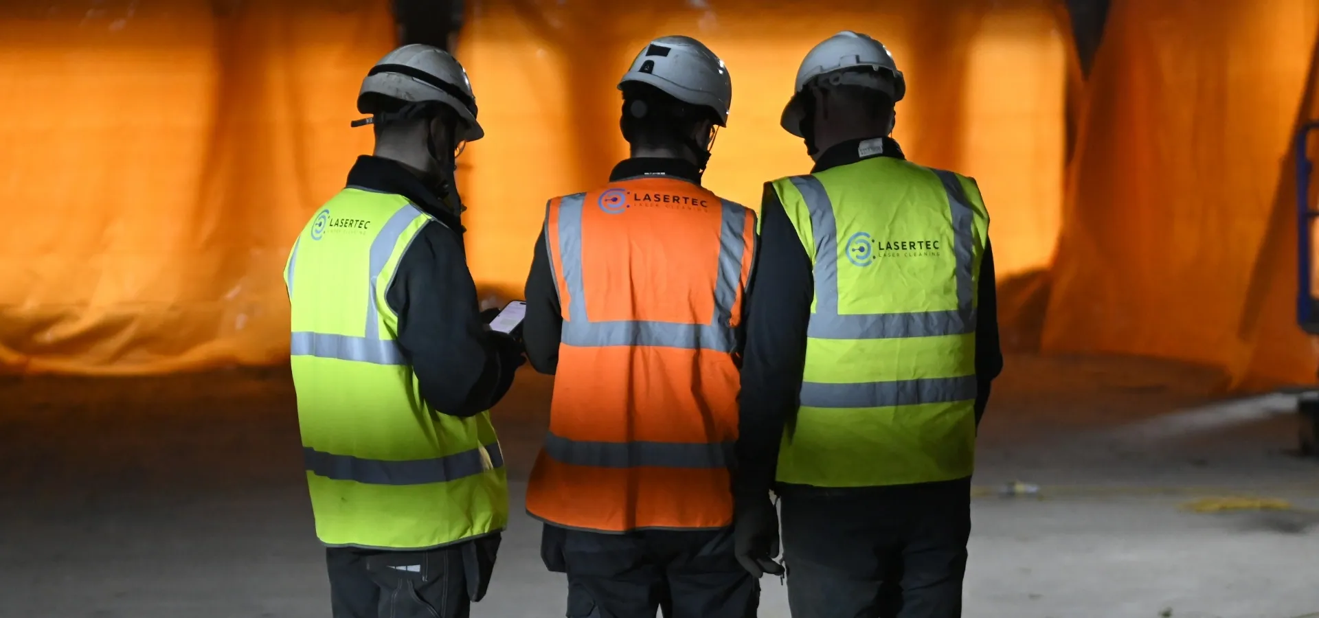 Three LaserTec team members in hi-vis and hard hats assessing a surface on site before laser cleaning work begins