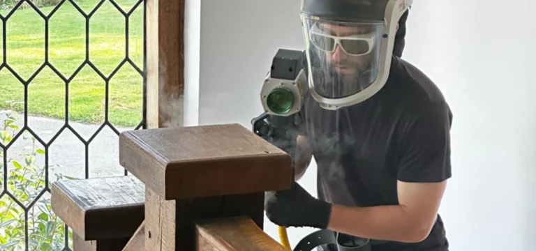 LaserTec technician in respirator hood and laser safety eyewear stripping varnish from an oak pew with a handheld laser