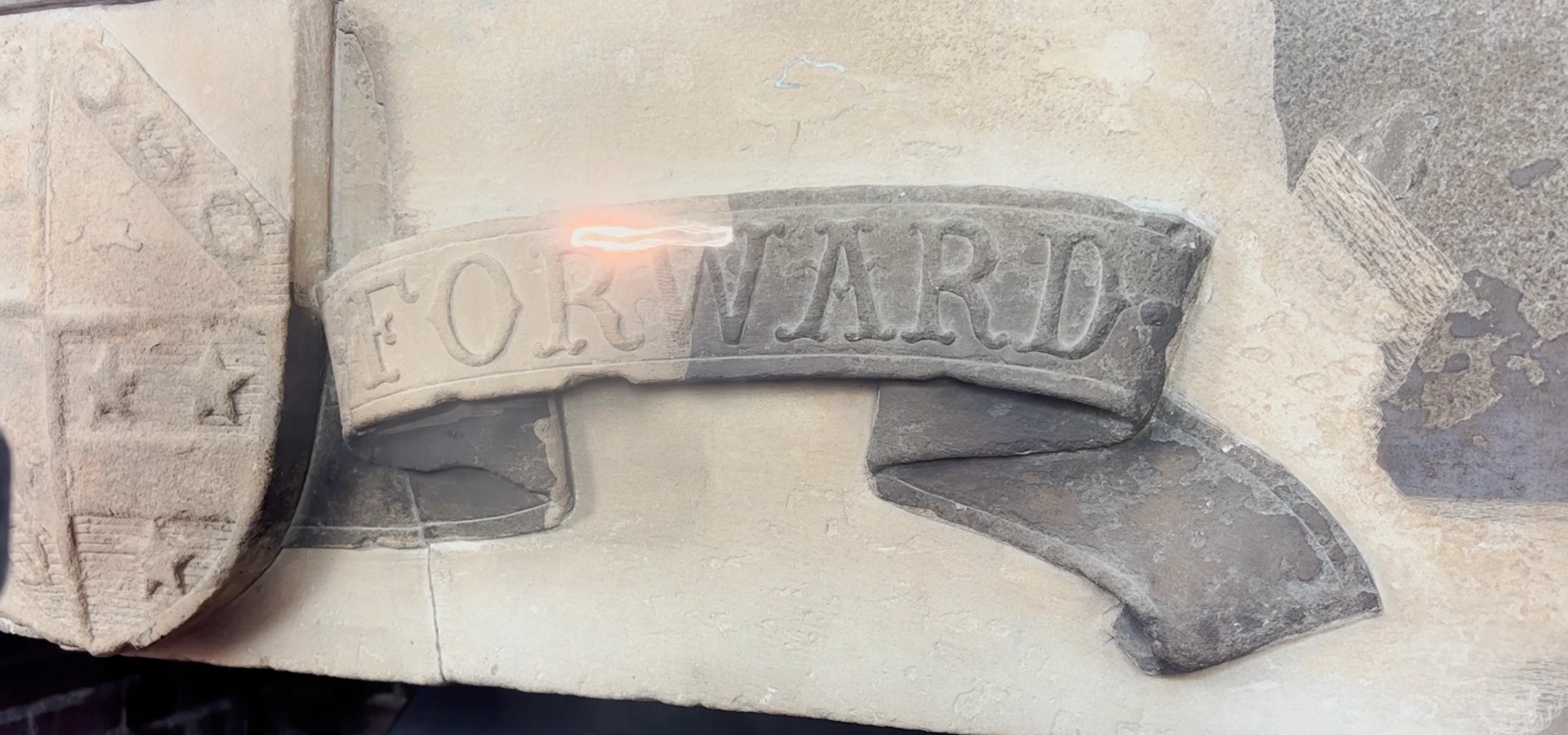 Laser cleaning in progress on a carved stone scroll bearing the word FORWARD, with the left half cleaned to pale stone and the right half still covered in dark soiling